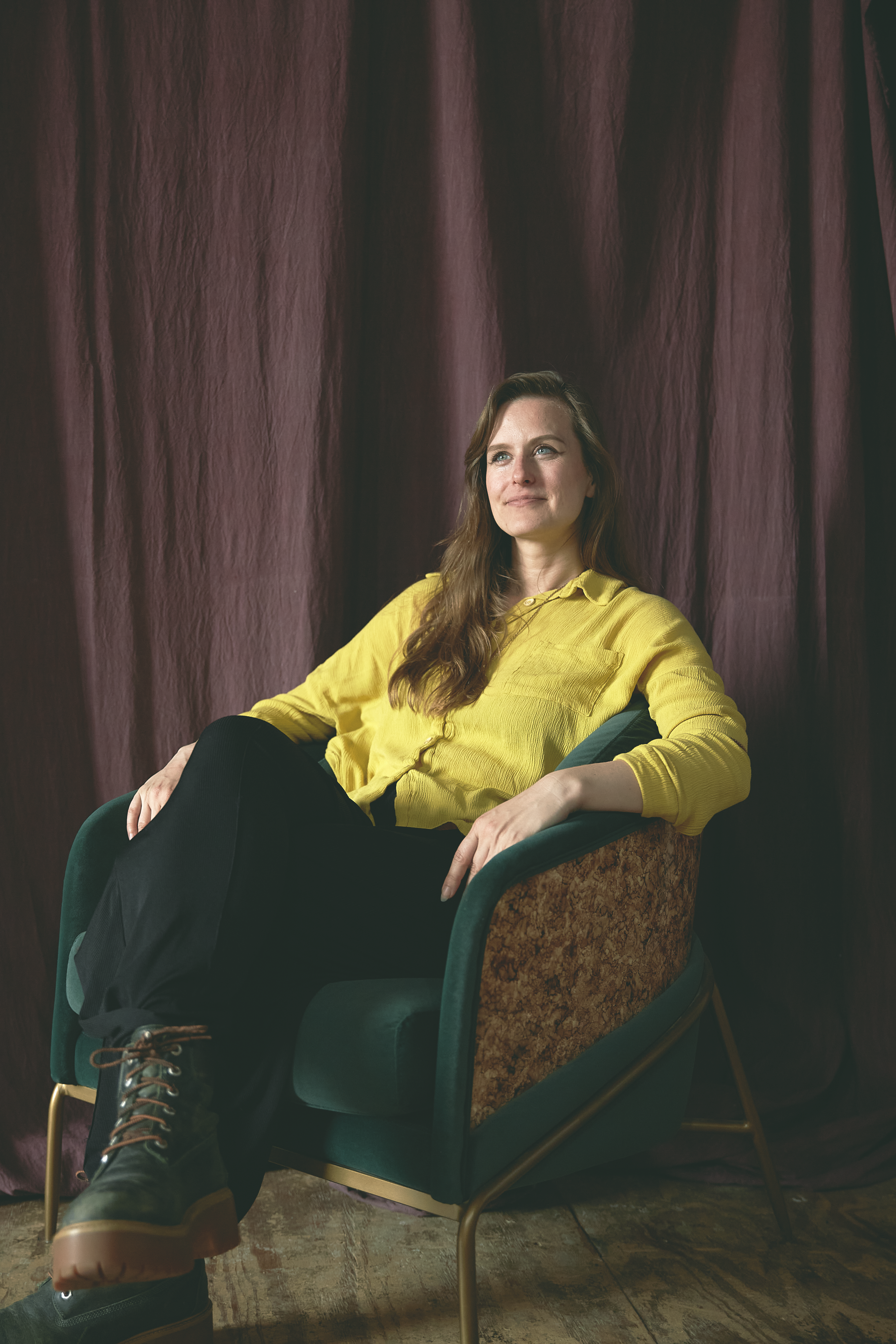 Viktoryia seated in a green velvet armchair in front of a plum curtain